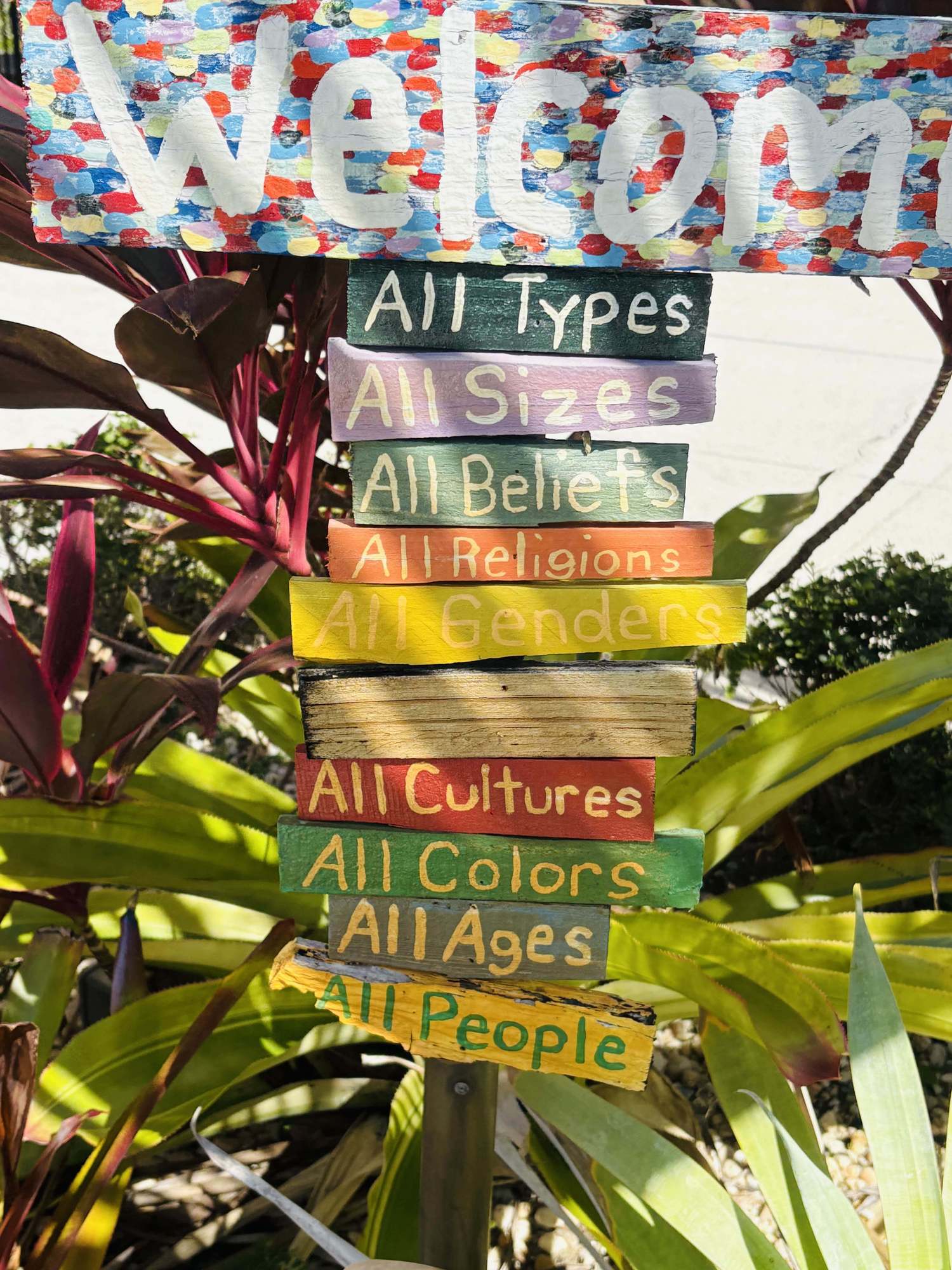 A colorful hand-painted garden sign reading: Welcome — all types, all sizes, all beliefs, all religions, all genders, all cultures, all colors, all ages, all people
